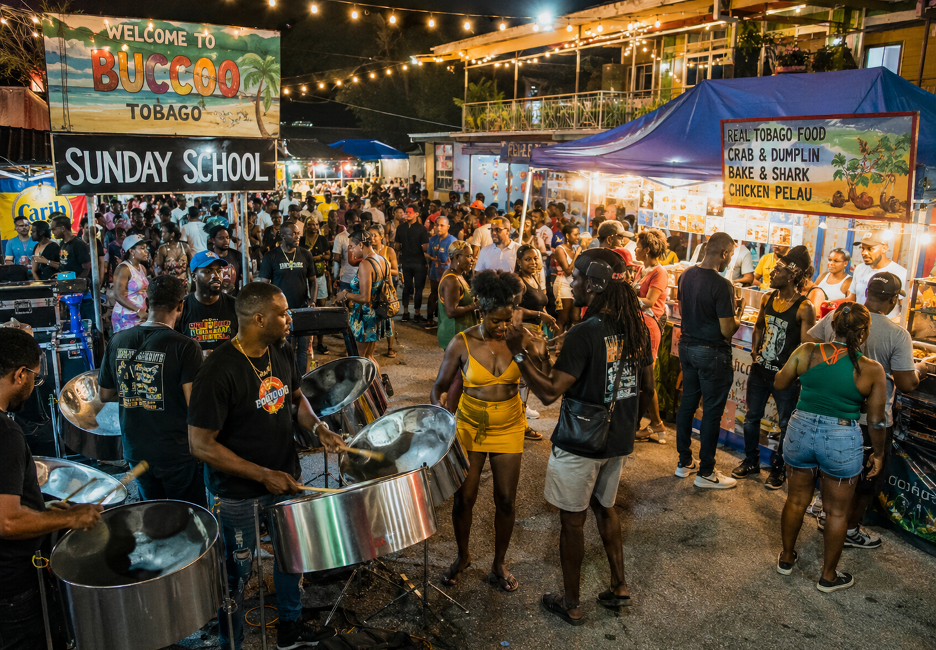 Night market scene in Buccoo, Tobago at night with a steel drum band performing and crowded food stalls under string lights.