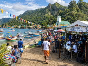 Crowd at Charlotteville Fisherman Festival by a harbor with colorful boats tents and a live band performing near the water and green hills