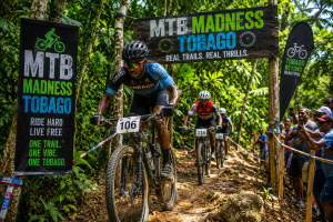 Leader on a mountain bike no 106 rides a muddy Tobago trail past banners reading MTB Madness Tobago as cheering spectators line the course