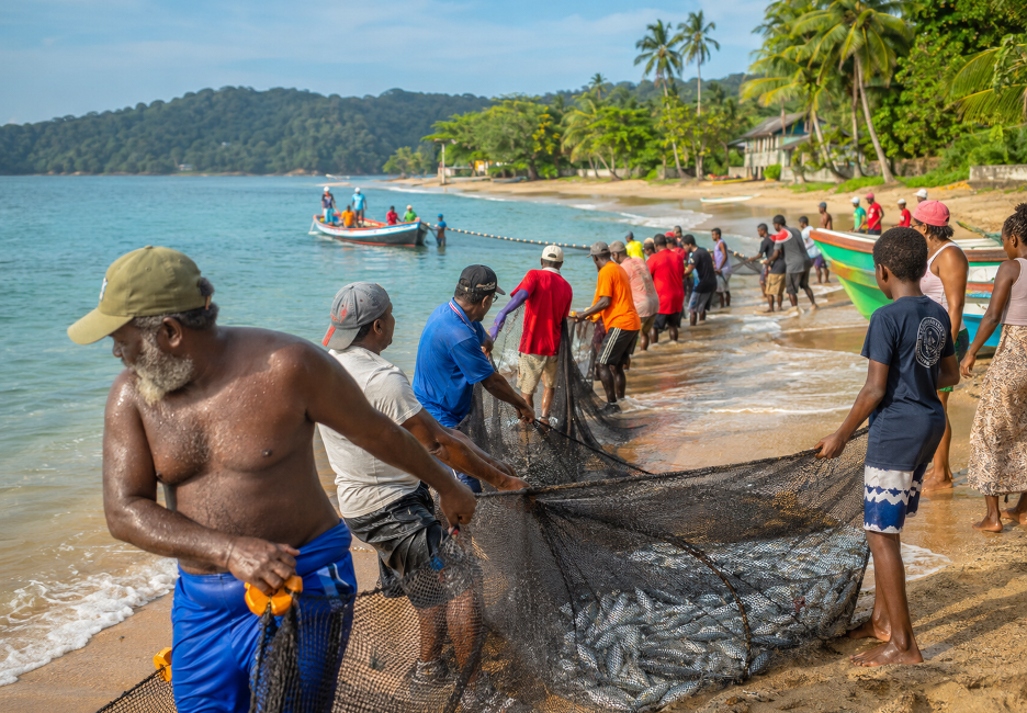 Group of fishermen hauling a large fishing net onto a sandy tropical beach as boats float nearby in calm water.