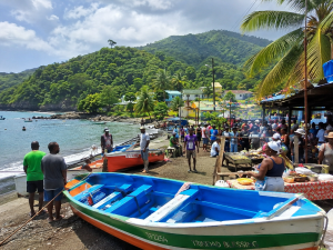 Bustling tropical beach market with colorful boats on shore and people gathered near food stalls against green hills and a blue sea