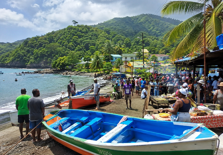 Bustling tropical beach market with colorful boats on shore and people gathered near food stalls against green hills and a blue sea.