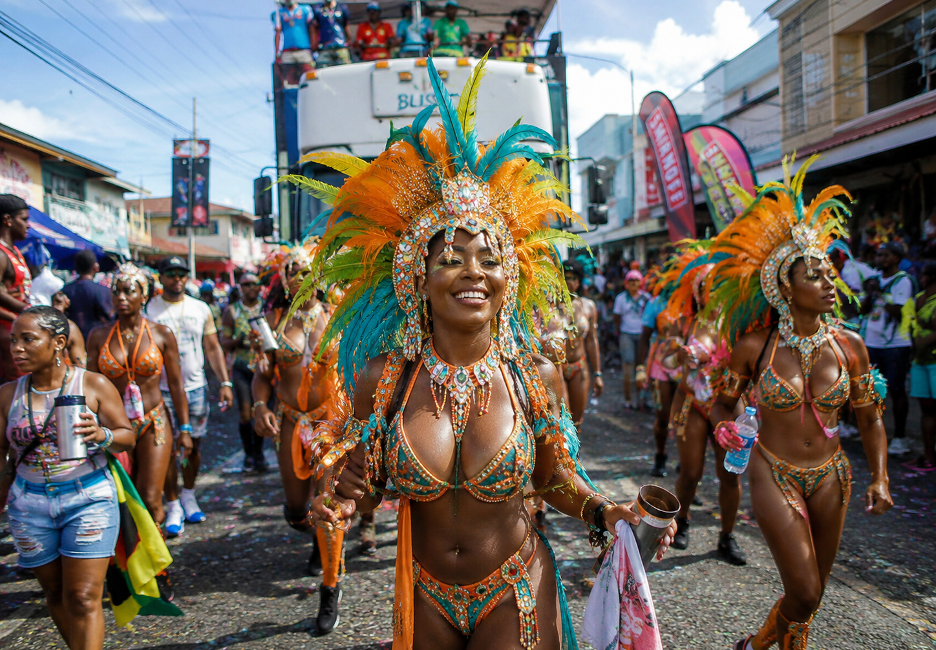 Carnival parade with dancers in bright turquoise and orange feather headdresses and sequined bikinis marching down a street to a cheering crowd.