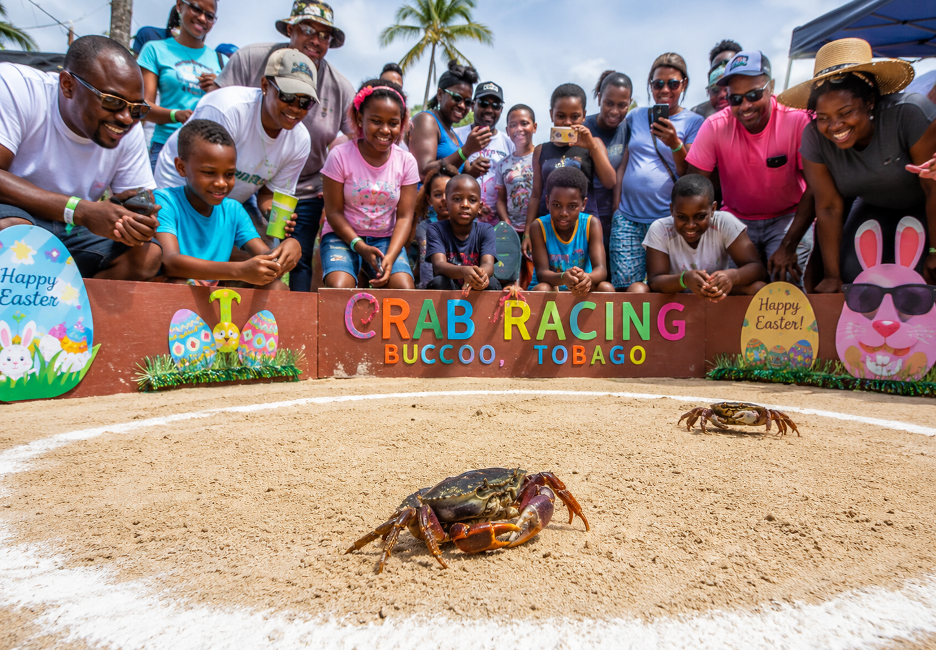 Diverse crowd watches a live crab race on a sandy track in Buccoo, Tobago, with a colorful 'CRAB RACING' sign and Easter decorations nearby.